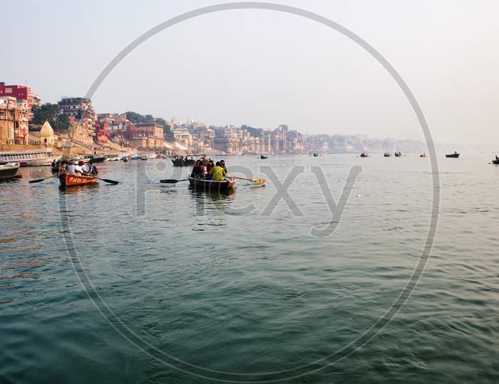 Varanasi, India - November 01, 2016: Tourist And Pilgrim Sightseeing On Wooden Boats In Ganges River Against Ghat And Banaras Cityscape During Foggy Weather.