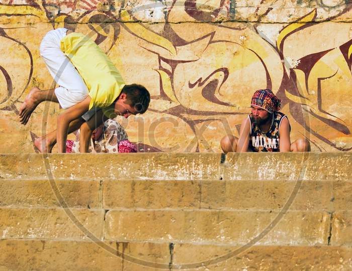 Varanasi, India - November 01, 2016: Couple Of Foreigner Men In Hipster Style Doing Yoga In The Morning With A Trainer Against Wall With Graffiti In The City Of Banaras Situated In Uttar Pradesh
