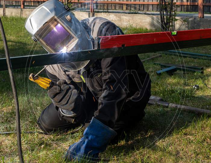 A Young  Man Welder In  Uniform, Welding Mask And Welders Leathers, Weld  Metal  With A Arc Welding Machine At The Construction Site  In Background  A Grass