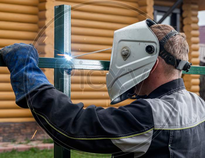 A Young  Man Welder In  Uniform, Welding Mask And Welders Leathers, Weld  Metal  With A Arc Welding Machine At The Construction Site  In Background  A Country House
