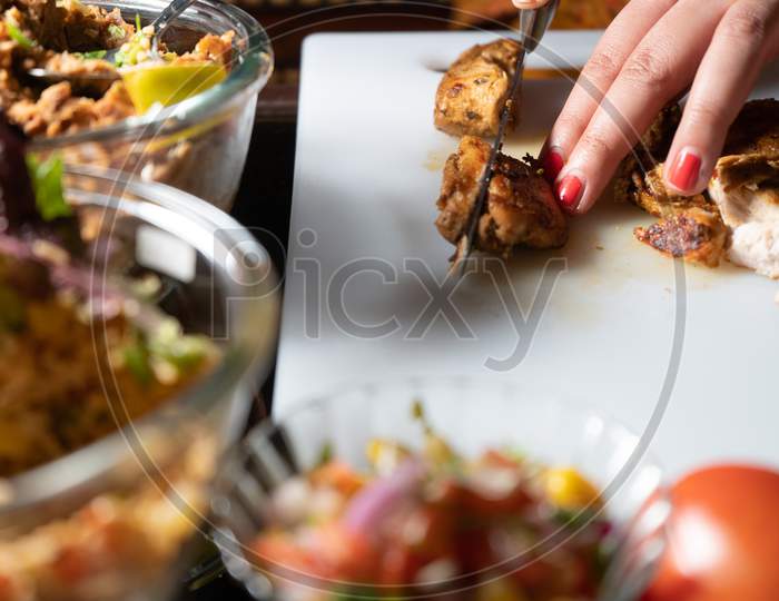 Woman Using A Sharp Knife To Cut Meat, Chiken, Mutton, Beef On A White Chopping Board With Fresh Vegetables Like Tomatoes, Rice And More To Prepare Indian Mexican Food At Home