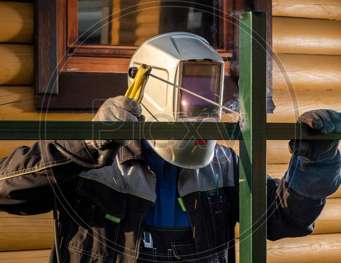Man Welder In Welding Mask, Building Uniform And  Protective Gloves Brews Metal Welding Machine On Street Construction, In The Background Country House In Summer Day