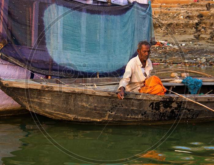Varanasi, India - November 01, 2016: An Old Man Sitting Alone On A Wooden Boat Fishing For Living Against A Cloth Being Dried In The City Of Banaras Located In Uttar Pradesh State.