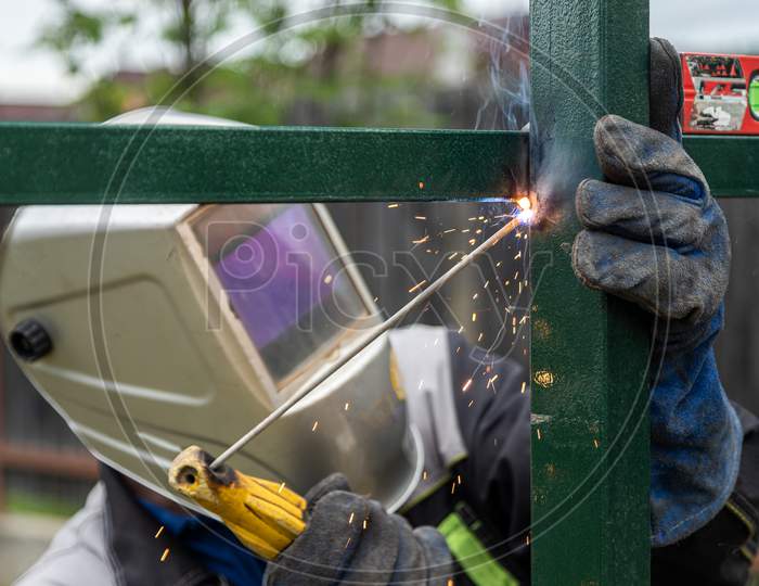 A Strong Man Is A Welder In A Welding Mask And Welders Leathers, A Metal Product Is Welded With A Welding Machine In The Outside, Yellow Sparks Fly To The Sides