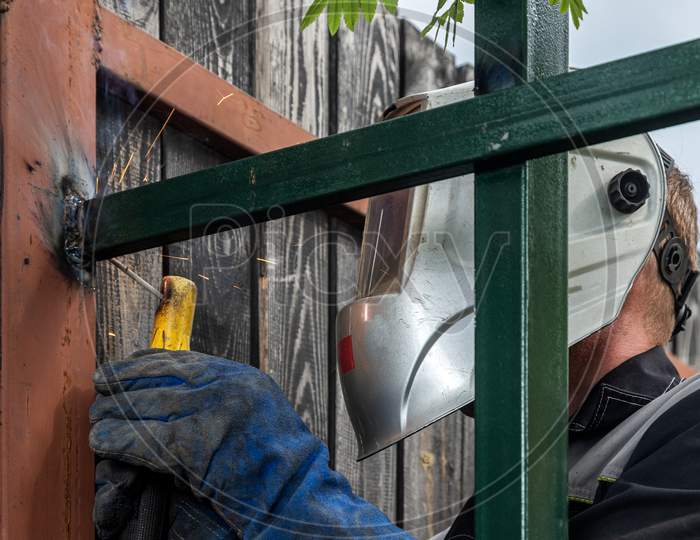 Man Welder In Welding Mask, Building Uniform And  Protective Gloves Brews Metal Welding Machine On Street Construction, In The Background Fence