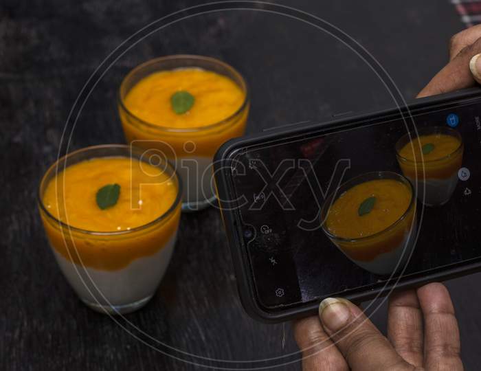 A Female Hand Taking Mobile Photo Of Two Glasses Of Delicious Mango Lassi Drink Decorated With Mint Leaves In Black Wooden Background.