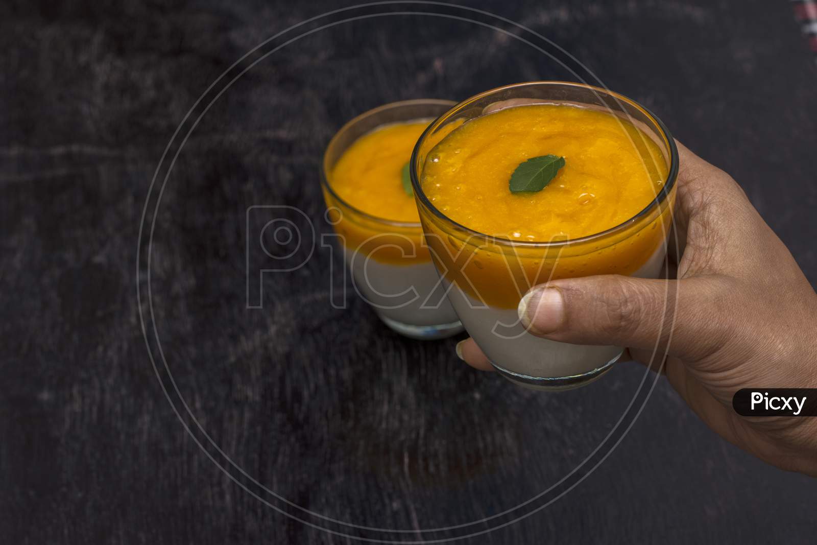 A Female Hand Taking On Of The Two Glasses Of Delicious Mango Lassi Drink Decorated With Mint Leaves In Black Wooden Background.