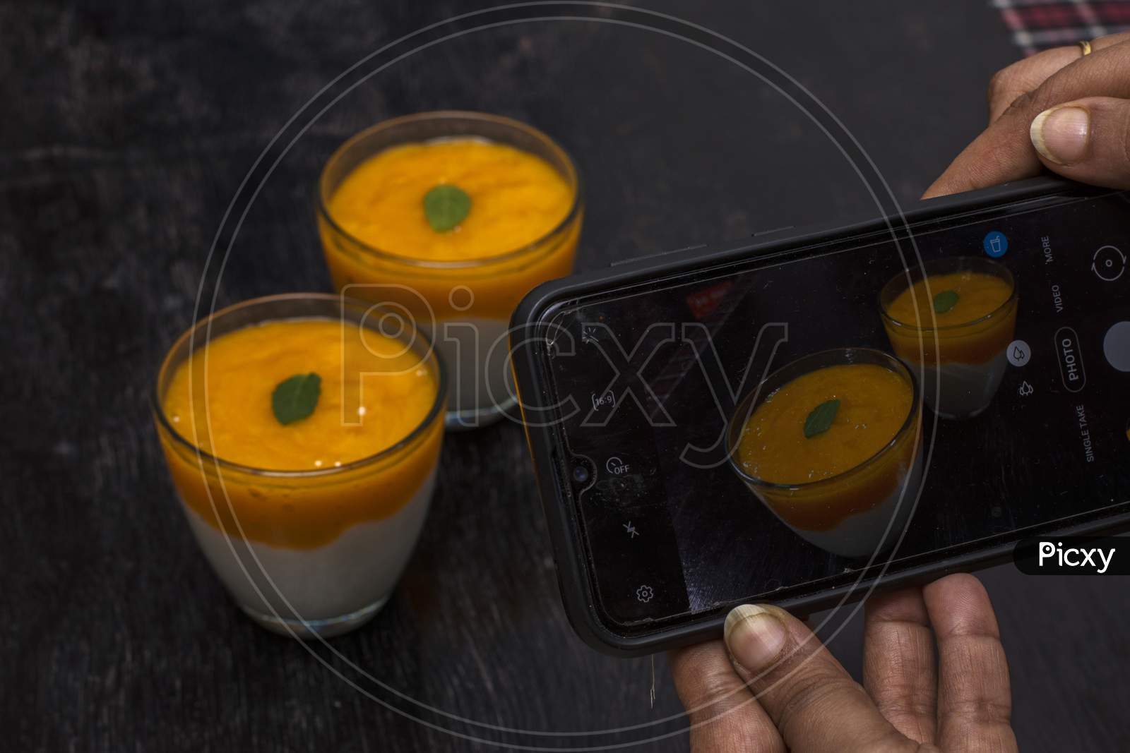 A Female Hand Taking Mobile Photo Of Two Glasses Of Delicious Mango Lassi Drink Decorated With Mint Leaves In Black Wooden Background.
