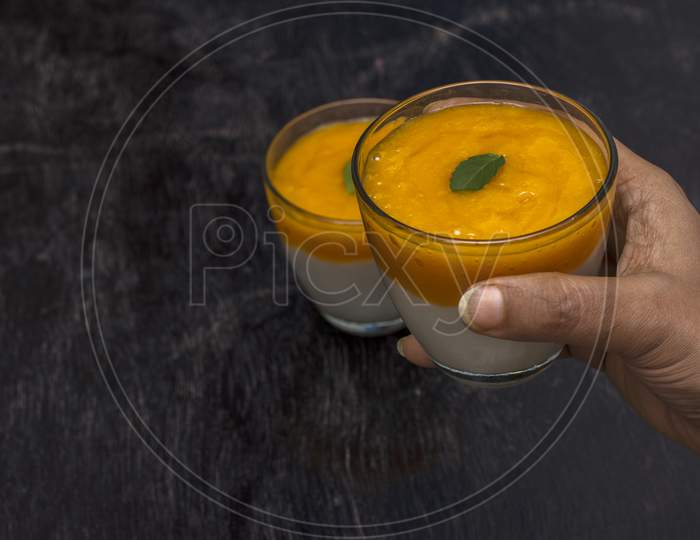 A Female Hand Taking On Of The Two Glasses Of Delicious Mango Lassi Drink Decorated With Mint Leaves In Black Wooden Background.