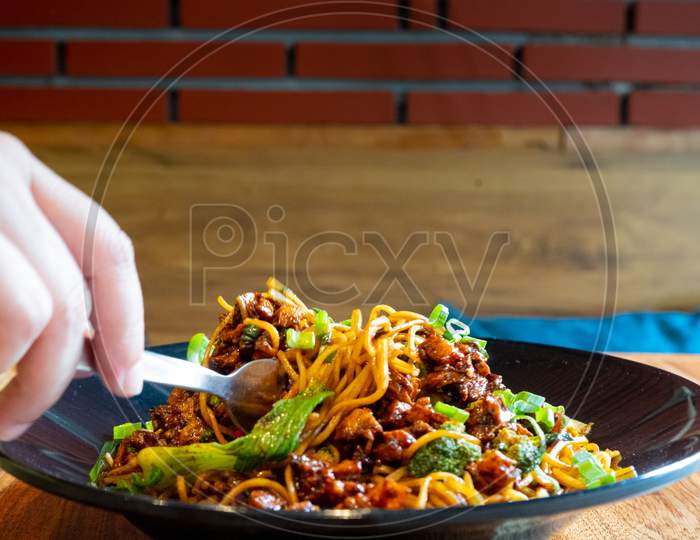 Woman With Fork Picking Up Indo Chinese Manchurian Hakka Street Food Noodles From A Black Bowl Placed On A Wooden Table In A Small Street Food Eatery While Travelling In Delhi