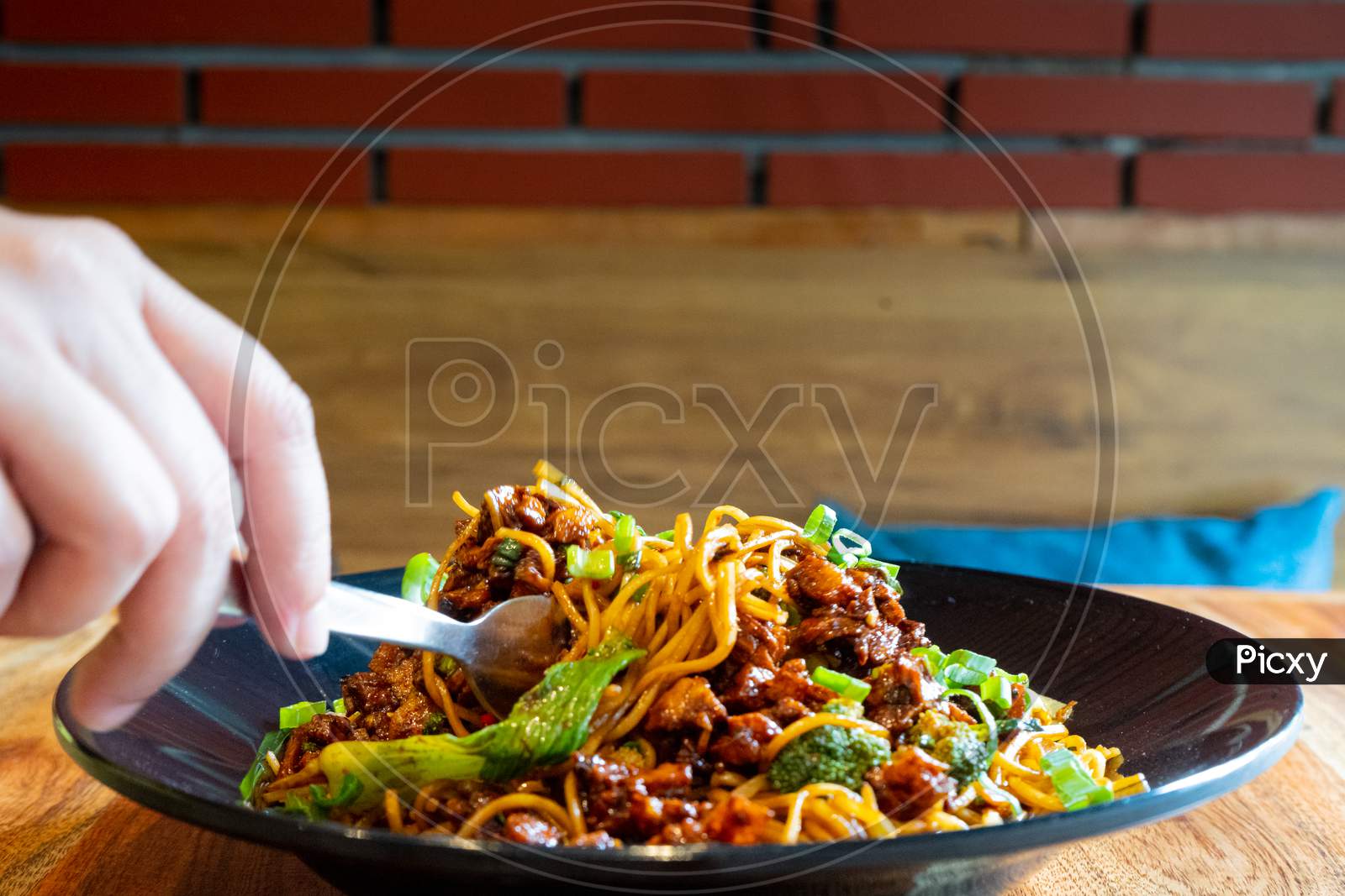 Woman With Fork Picking Up Indo Chinese Manchurian Hakka Street Food Noodles From A Black Bowl Placed On A Wooden Table In A Small Street Food Eatery While Travelling In Delhi