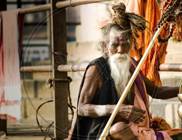 Varanasi, India - November 01, 2016: Portrait Of A Hindu White Bearded Sadhu, Pilgrim Or Aghori Baba With A Lota, Braided Necklace And Dreadlock Against Ganges River Ghat During Day.