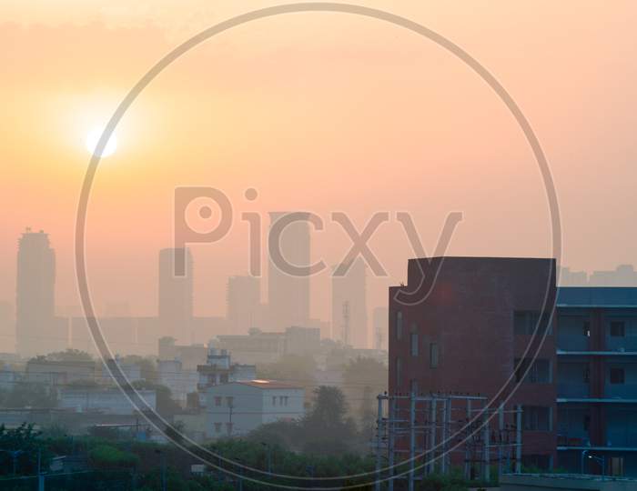 Wide Shot Of Gurgaon Delhi Cityscape Showing A Brick Building And Houses In The Foreground And Tall Buildings With Many Floors In The Fog Haze Obsured Distance Against The Sun Rising