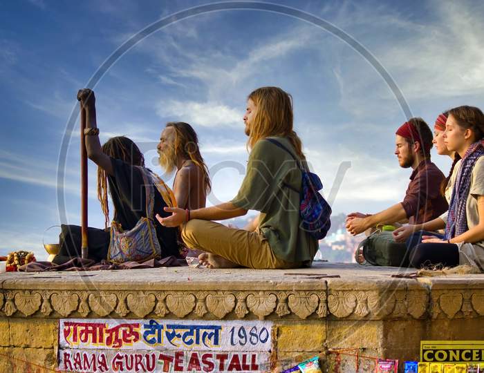 Varanasi, India - November 01, 2016: Bunch Of Tourists, Sadhu Baba And Pilgrims In Sitting Posture Meditating Together On A Pile Of Rock During Morning Facing Ganges River And Sunrise. Uttar Pradesh