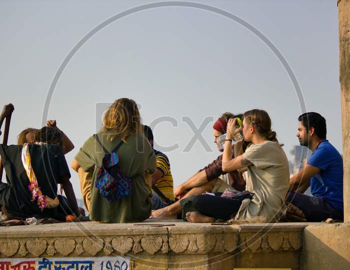 Varanasi, India - November 01, 2016: Bunch Of Tourists, Sadhu Baba And Pilgrims In Sitting Posture Meditating Together On A Pile Of Rock During Morning Facing Ganges River And Sunrise. Uttar Pradesh