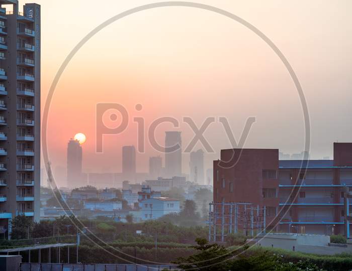 Dawn Shot Of Skyscraper And Small Houses In The Background With Tall Buildings With Homes Offices Hidden By Fog In The Background With The Sun Rising Behind Them