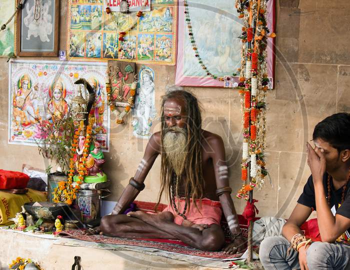 Varanasi, India - November 01, 2016: Portrait Of A Hindu White Bearded Sadhu, Pilgrim Or Aghori Baba With Closed Eyes Meditating. Dreadlocked Hindu Man With God Pictures On Ganges River Ghat.