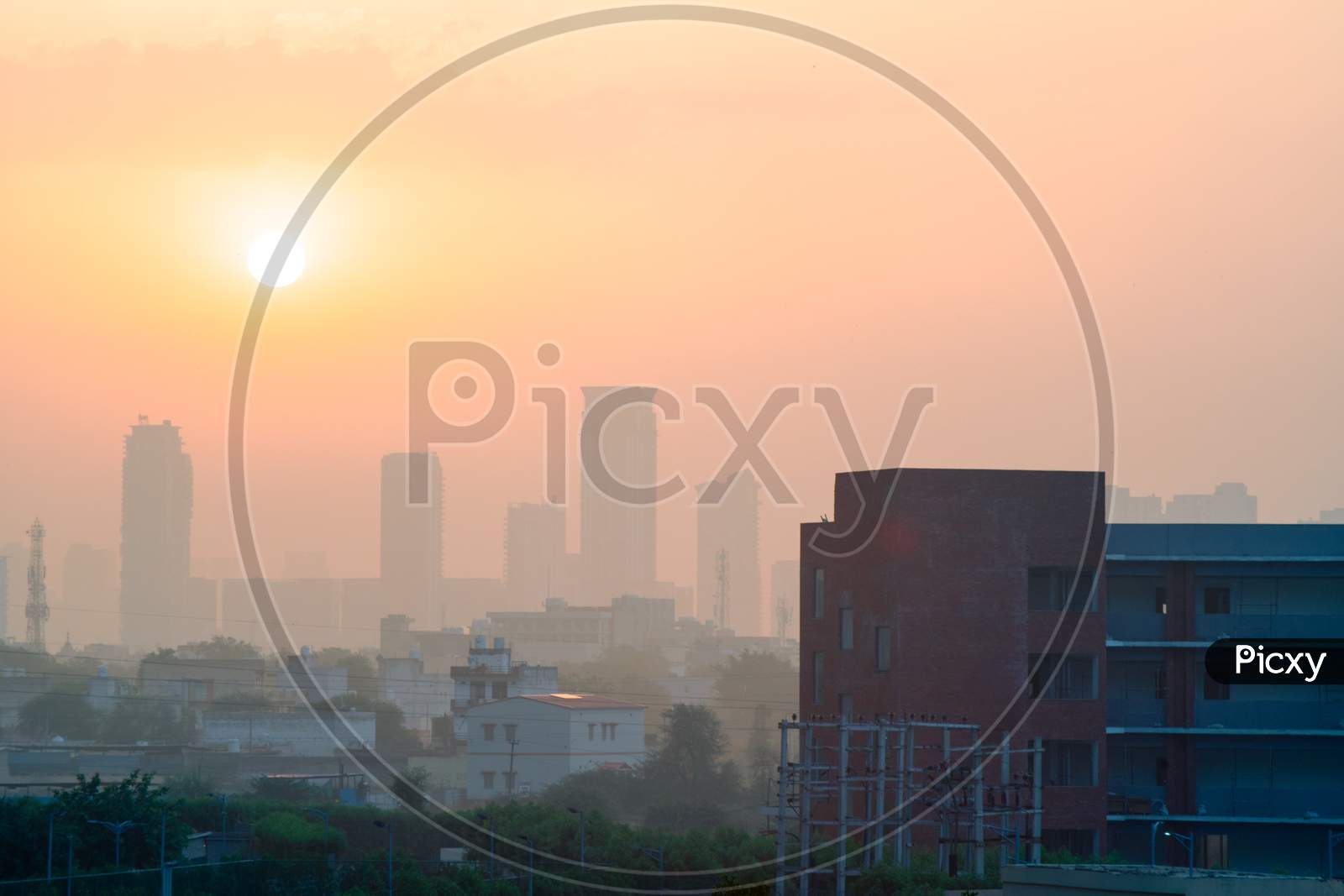 Wide Shot Of Gurgaon Delhi Cityscape Showing A Brick Building And Houses In The Foreground And Tall Buildings With Many Floors In The Fog Haze Obsured Distance Against The Sun Rising