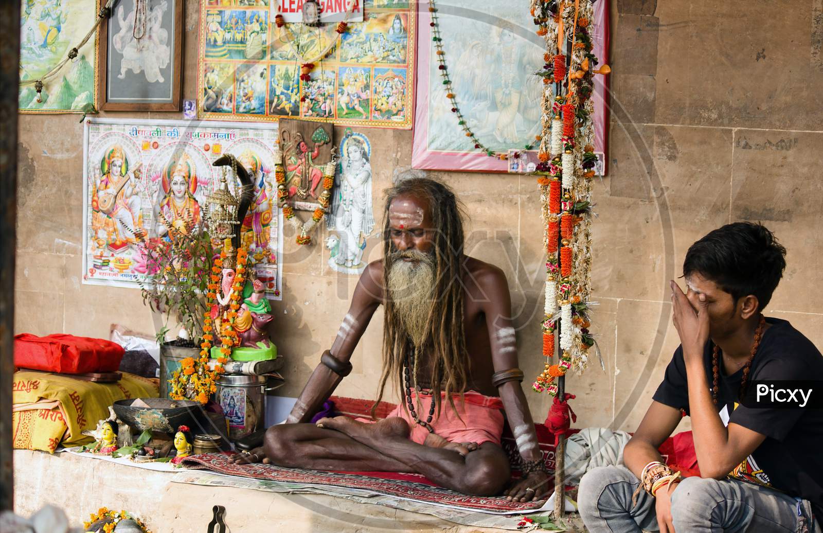 Varanasi, India - November 01, 2016: Portrait Of A Hindu White Bearded Sadhu, Pilgrim Or Aghori Baba With Closed Eyes Meditating. Dreadlocked Hindu Man With God Pictures On Ganges River Ghat.