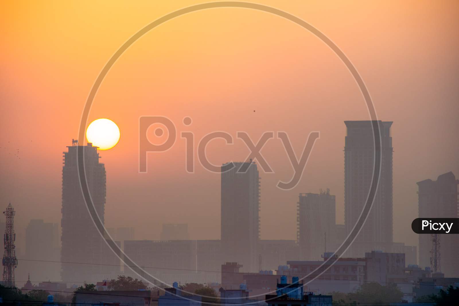 Dusk Dawn Shot Of Skyscrapers With Fog Around Them And The Sun Rising Behind Them Shot In Gurgaon Delhi India With Small Houses In The Foreground