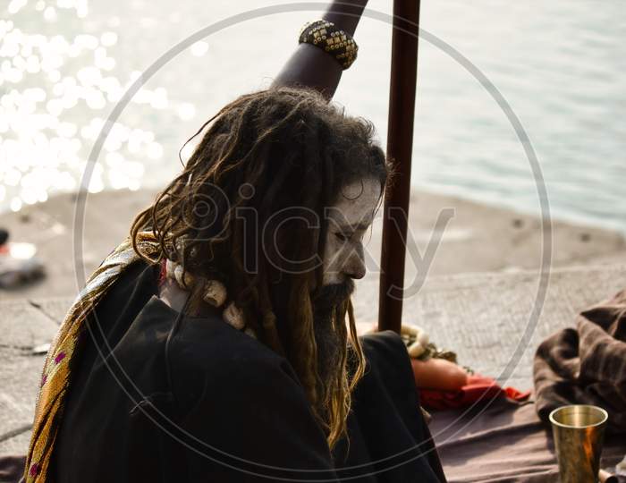 Varanasi, India - November 01, 2016: Side Profile Of A Hindu Bearded Sadhu, Pilgrim Or Aghori Baba With Closed Eyes Sitting With A Stick Against Ganges River During Day.