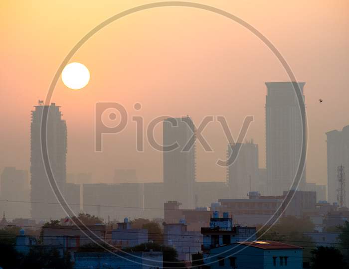 Dusk Dawn Shot Of Skyscrapers With Fog Around Them And The Sun Rising Behind Them Shot In Gurgaon Delhi India With Small Houses In The Foreground