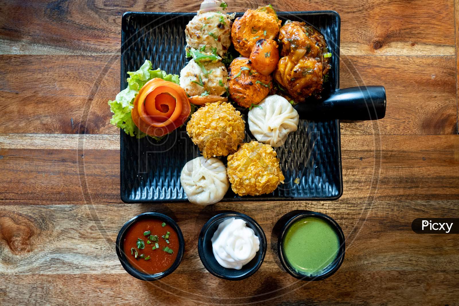 Top Down Flatlay Shot Of A Momo Platter With Crispy Tandoori Fried Momos Steamed Momos And Chicken Pakora With Green Sauce, Tartar And Soya On Wooden Table