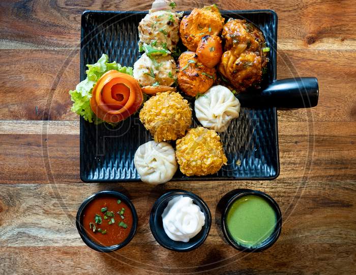 Top Down Flatlay Shot Of A Momo Platter With Crispy Tandoori Fried Momos Steamed Momos And Chicken Pakora With Green Sauce, Tartar And Soya On Wooden Table