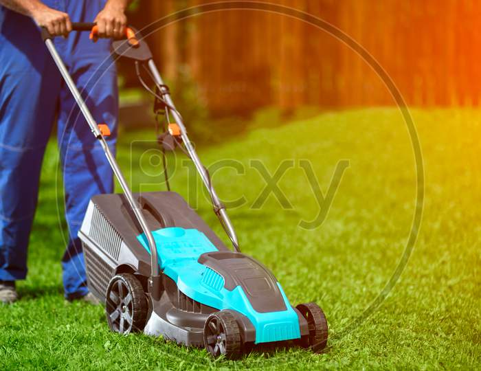 Close-Up Of A Man In Overalls With A Lawn Mower Cutting Green Grass In A Modern Garden. Lawn Mowing Machine.