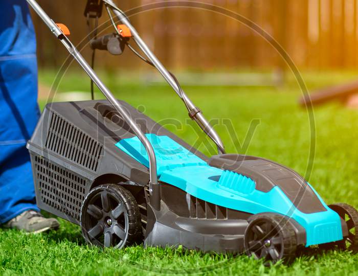 Male Worker On The Street Working On Mowing The Lawn With The Help Of A Modern Lawn Mower Near A Country House