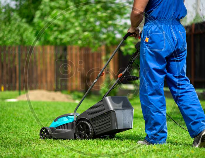 A Man With An Electric Lawn Mower Mowing The Lawn.Adult Man Pruning And Landscaping A Garden, Mowing Grass, Lawn, Paths.