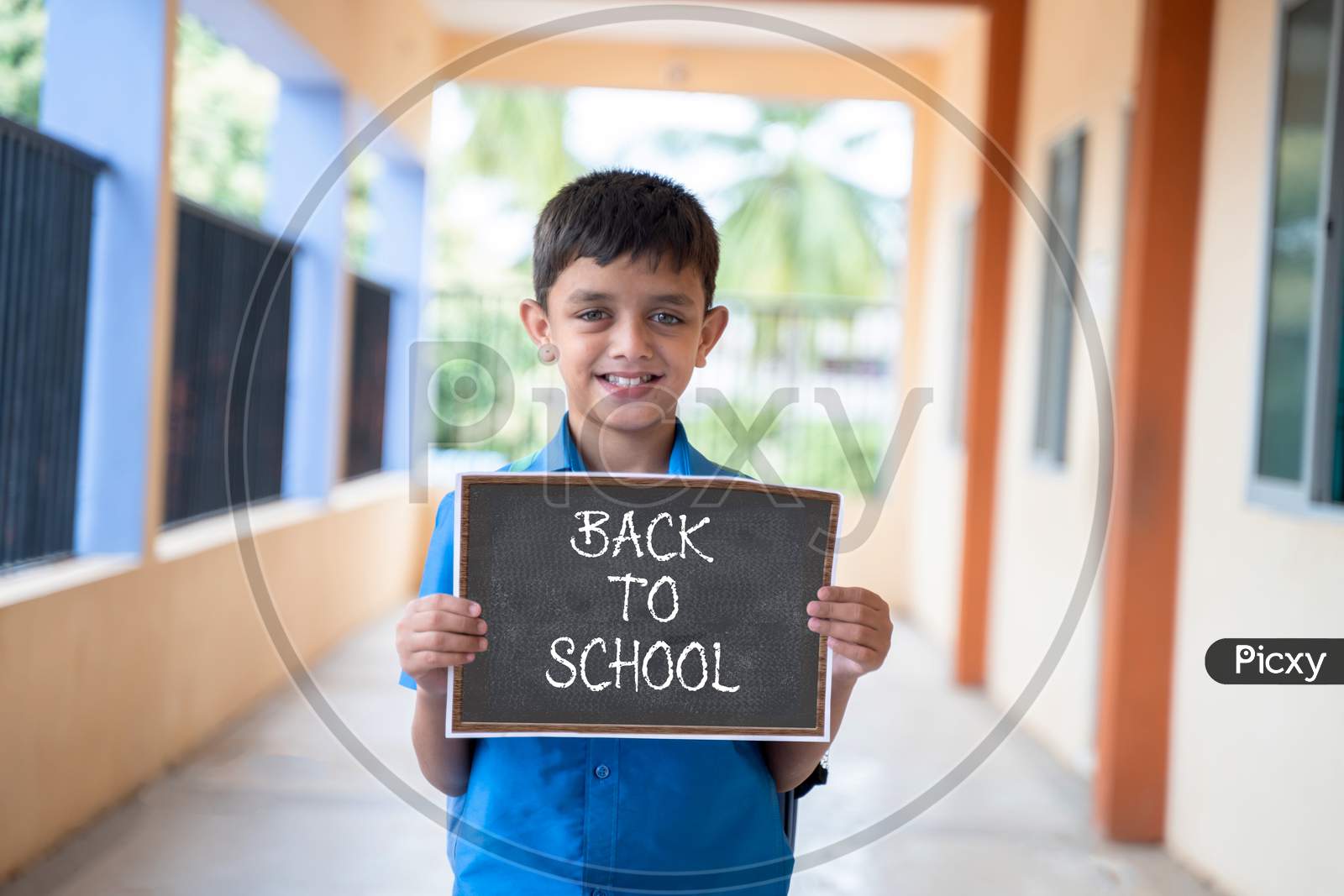 Smiling Young Kid Looking Camera And Showing By Back To School Sign Board School At Corridor - Concept Of School Reopen And Back To School.