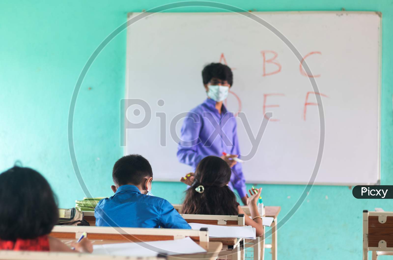 Focus On Middle Kid,Teacher In Medical Mask With Limited Kids Teaching At Classroom With Coronavirus Covid-19 Safety Measures At Classroom - Concept Of New Normal, Education And Back To School.