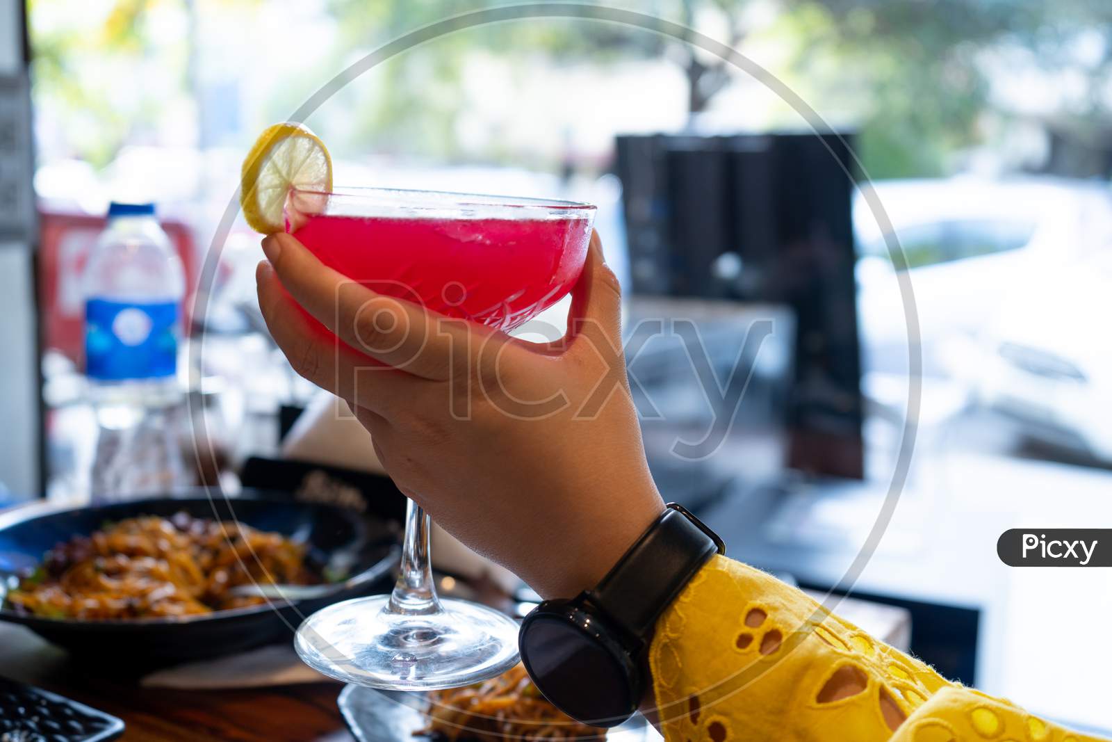 Young Indian Woman In Yellow Dress Watch Holding A Large Wine Glass Filled With A Red Cocktail Mocktail Of Strawberry Cherry With A Lemon Slice