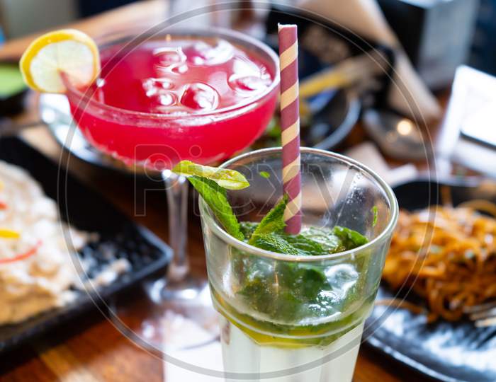 Close Up Shot Of Red Pink Colored Drink Made Of Strawberry Cherry And Berries And Mojito With A Lime Slice And Ice Placed On A Table With Out Of Focus Food In A Trendy Upmarket Restaurant In India