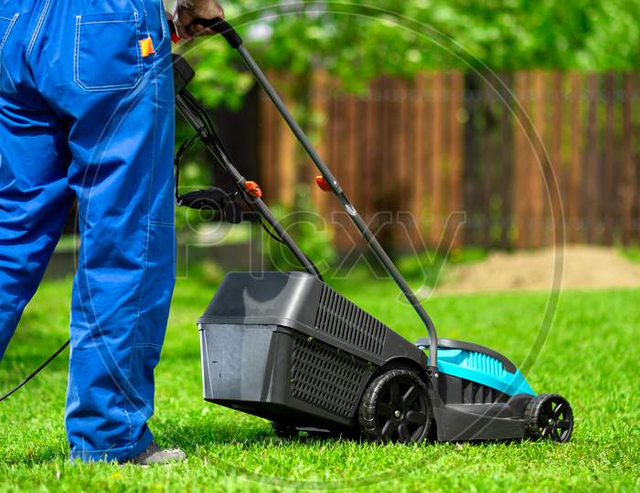 Close-Up Of A Man In Overalls With A Lawn Mower Cutting Green Grass In A Modern Garden. Lawn Mowing Machine.