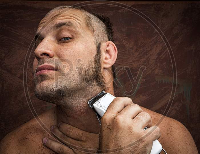Close-Up Portrait Of Handsome Shirtless Man Shaving His Beard With An Electric Razor   Against Brutal Background. Concept Of Male Home Care Without Beauty Salons
