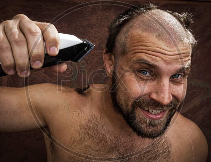 Close-Up Portrait Of Handsome Shirtless Man Shaving His Head With An Electric Razor And Gritting His Teeth, Against Brutal Background. Concept Of Male Home Care Without Beauty Salons