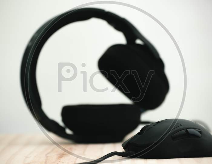 Headphone And The Mouse In A Wooden Computer Table, Mouse In Focus As Headset Blurred Behind, Darker Objects Against The Lighter Background Photograph.