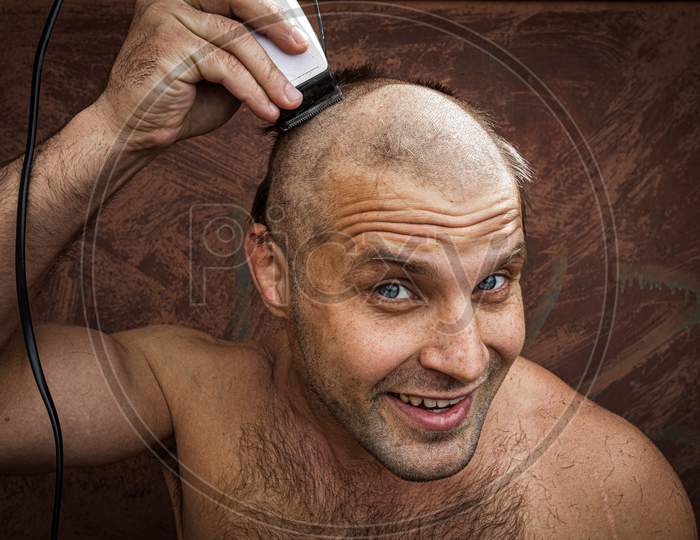Close-Up Portrait Of Handsome Shirtless Man Shaving His Head With An Electric Razor And Smilling, Against Brutal Background. Concept Of Male Home Care Without Beauty Salons