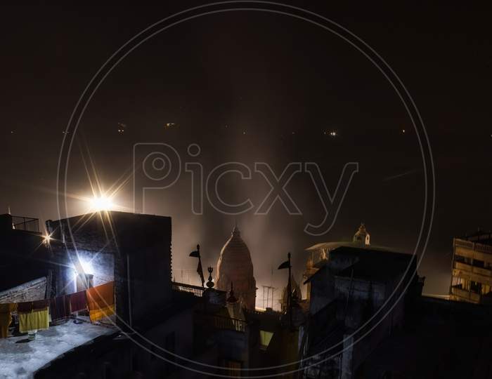 Night View Showing Rooftop Of Random Houses With Clothes Drying On Rope Against Temple Top With Smokes Rising Due To Aarti And Barely Visible Ganges River Int He Background
