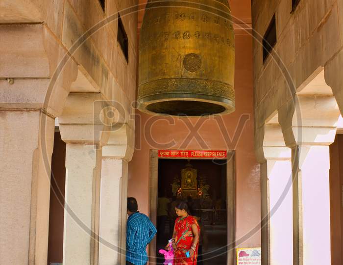 Varanasi, India - November 02, 2016: A Woman In Saree Visiting A Buddhist Temple Mulagandhakuti Vihara In Sarnath, Site Of Buddha`S First Sermon. Interior Of Temple Showing A Sacred Giant Prayer Bell