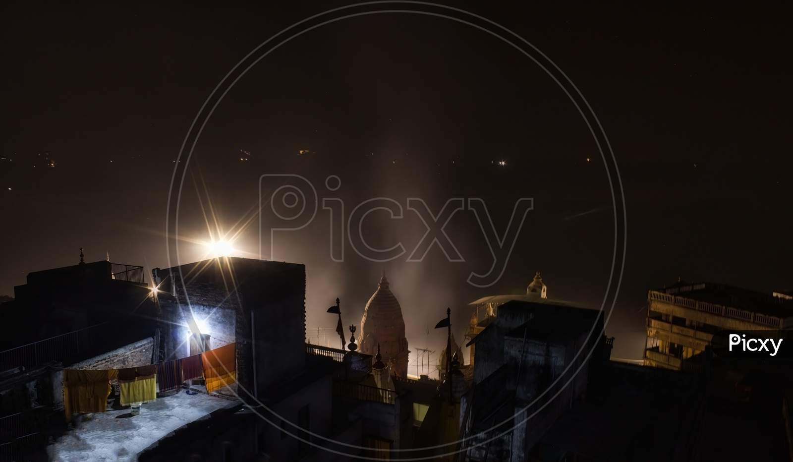 Night View Showing Rooftop Of Random Houses With Clothes Drying On Rope Against Temple Top With Smokes Rising Due To Aarti And Barely Visible Ganges River Int He Background