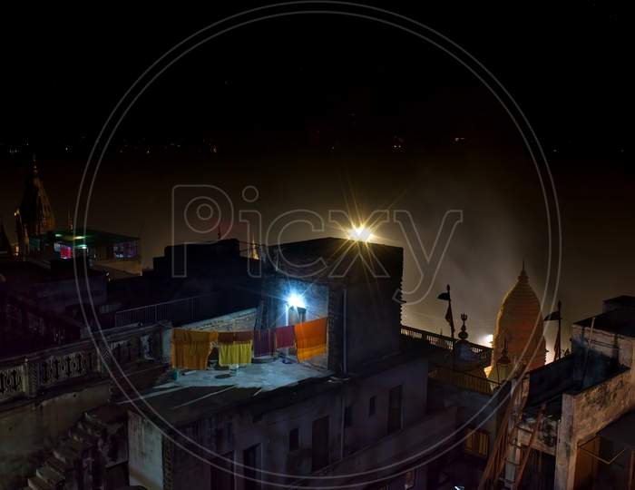 Wide Angle Night View Showing Rooftop Of Random Houses With Clothes Drying On Rope Against Temple Top With Smokes Rising Due To Aarti And Barely Visible Ganges River In The Background
