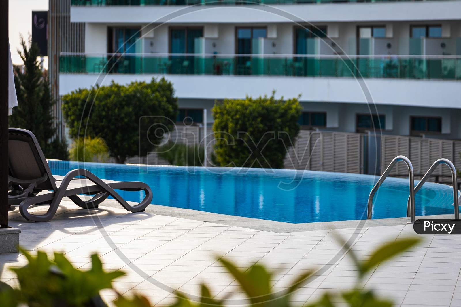 A Comfortable Gray Sun Lounger Set Against The Backdrop Of A Beautiful Transparent Pool And Hotel. The Concept Of A Relaxing Seaside Vacation For Two