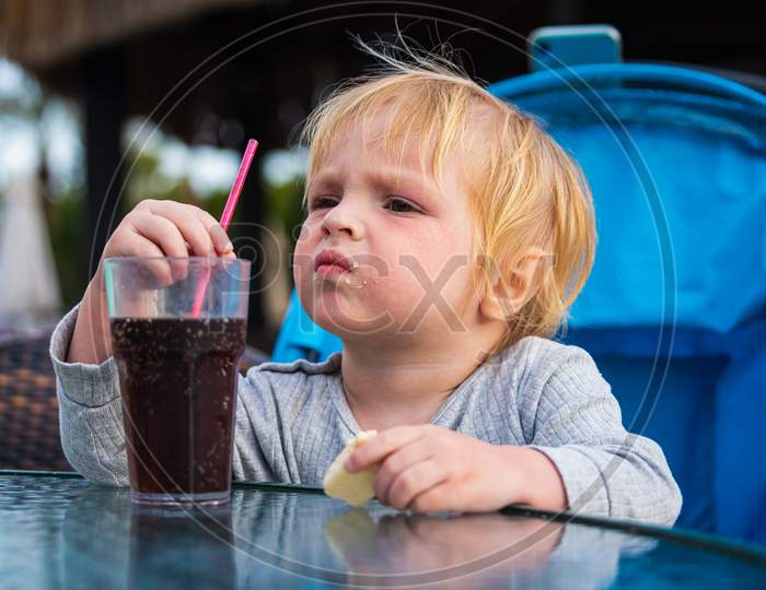 A Little Boy Is Sitting On A Chair And Happily Drinks Sparkling Water From A Glass Through A Straw. Feeding Process. The Baby Is Learning To Eat. Baby Food, Family, Child, Food