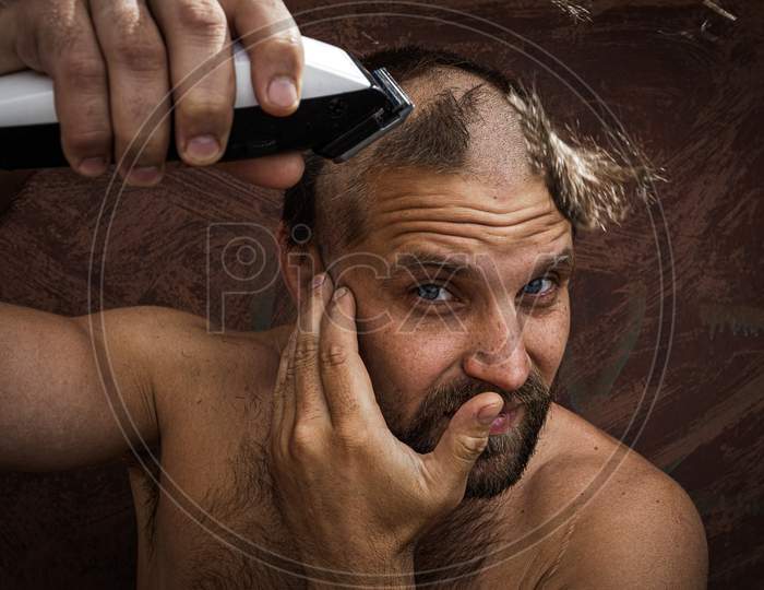 Caucasian Man Trying To Shave His Head With An Electric Razor. A Brutal Bald Man Holds A Razor In His Hand And Shaves Stubble On A Metal Background