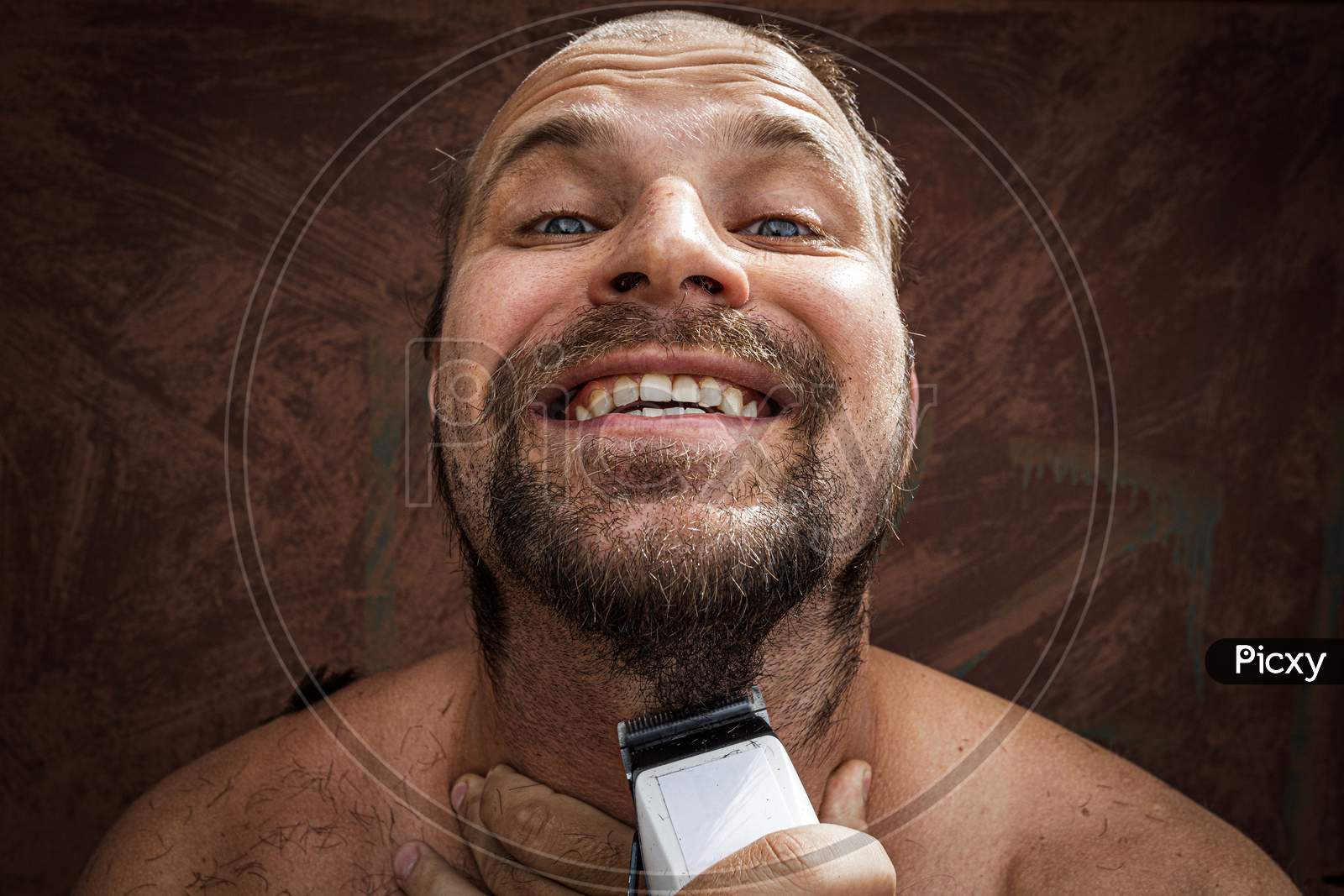 Close-Up Portrait Of Handsome Shirtless Man Shaving His Beard With An Electric Razor And Gritting His Teeth, Against Brutal Background. Concept Of Male Home Care Without Beauty Salons
