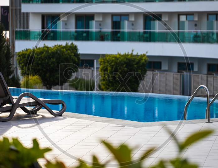 A Comfortable Gray Sun Lounger Set Against The Backdrop Of A Beautiful Transparent Pool And Hotel. The Concept Of A Relaxing Seaside Vacation For Two