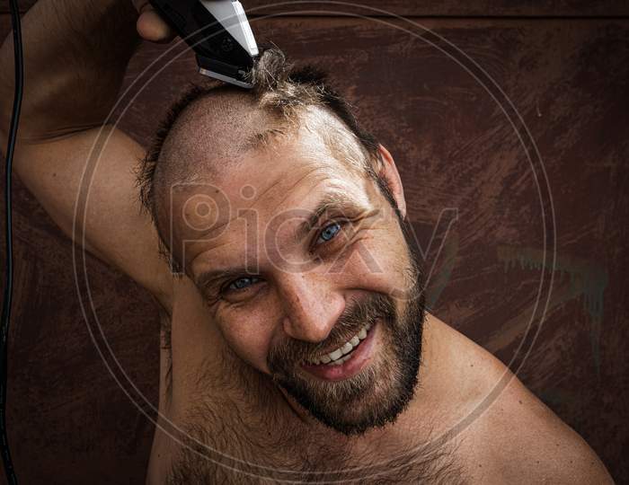 An Adult Cheerful Man With A Beard Shaves His Head At Home. The Guy Looks In The Bathroom Mirror And Uses The Electric Trimmer. Front View
